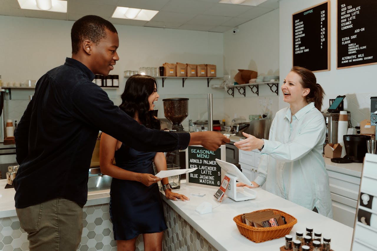 Customers and barista laughing and exchanging payment at a coffee shop counter.